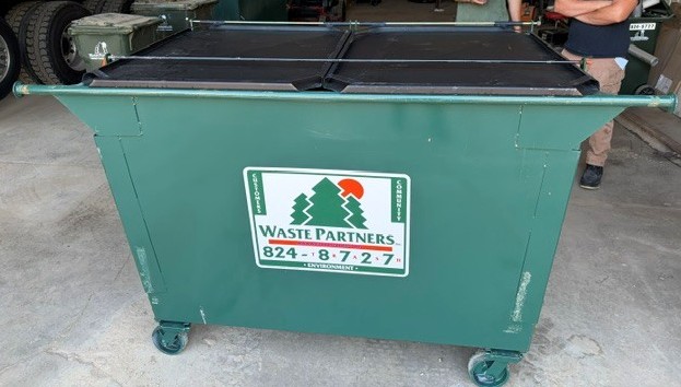 Green wheeled dumpster with a black lid, displaying a Waste Partners logo featuring trees and a red sun, along with a contact number.