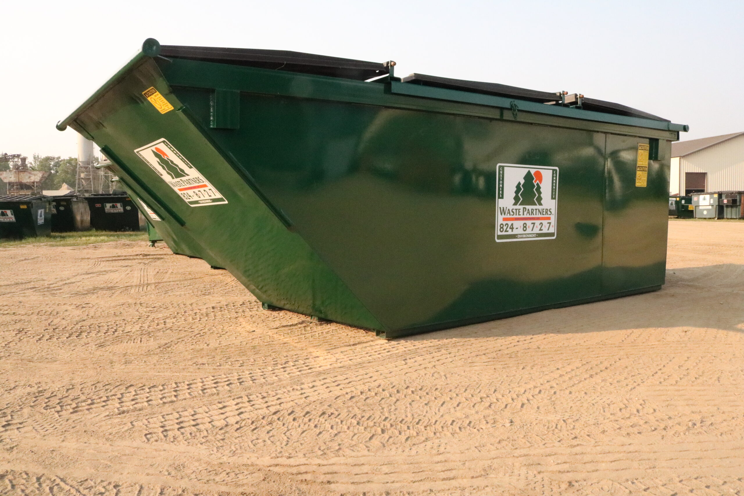 Large green industrial dumpster with warning labels parked on a dirt lot near buildings in an outdoor yard.